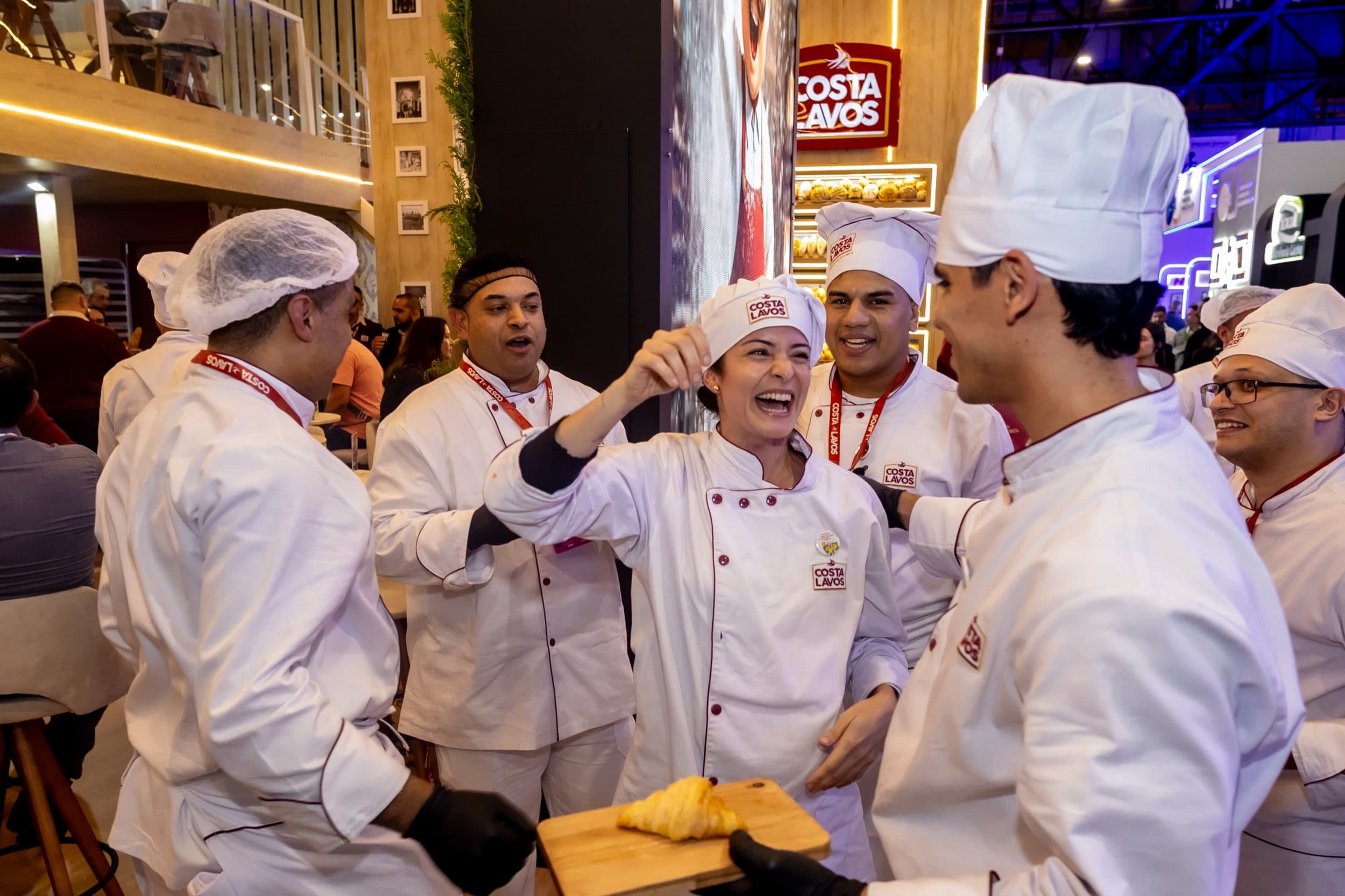 Profissionais de dólmã branco e touca com a marca Costa Lavos sorrindo juntos; um deles segura uma tábua com croissant.